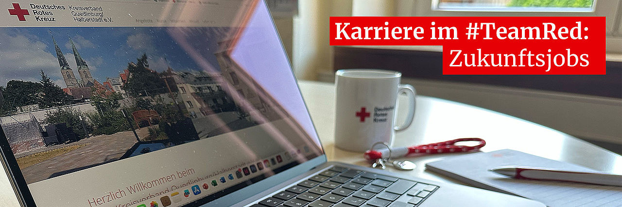 Laptop mit geöffneter DRK-Website auf Schreibtisch, daneben ein DRK-Schreibblock,-Kugelschreiber und Tasse